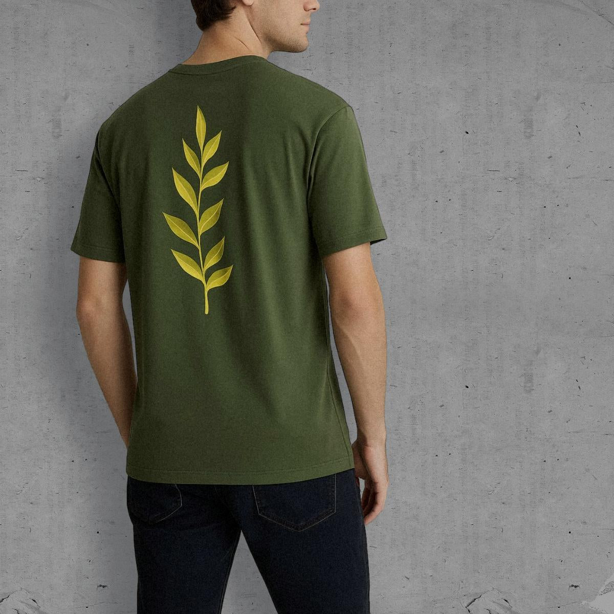 Person wearing a green t-shirt with a yellow leaf design on the back against a gray concrete wall.